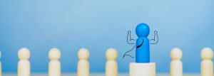 Images shows a row of wooden pins. A blue pin stands on a platform above the rest, with strong arms, a cape and a tie