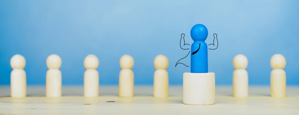 Images shows a row of wooden pins. A blue pin stands on a platform above the rest, with strong arms, a cape and a tie