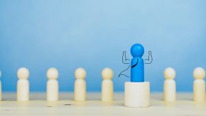 Image shows a line of wooden pins, with one blue pin standing above the rest on a platform. The blue pin has strong arms, a tie and a cape.