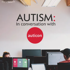 Office scene with two people working at computer desks. On the wall behind them is the text: 'AUTISM: In conversation with auticon,' with the word 'auticon' inside a red circle. The environment suggests a professional setting focused on autism-related discussions or initiatives by Auticon, a company known for employing autistic professionals in tech roles