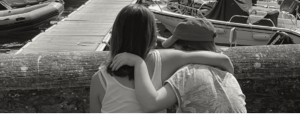 Holly and her sister have their arms around each other and look out towards a pier and fishing boats
