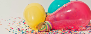 Three balloons (yellow, red and cyan) rest on the floor in a pile of confetti
