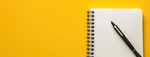 a black pen sits on the open page of a spiral bound note book, against a warm yellow background