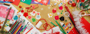 Flat lay of colorful arts and crafts supplies on a glittery gold background, including paintbrushes, colored pencils, glue, scissors, festive stickers, beads, string, and snowflake-themed paper.