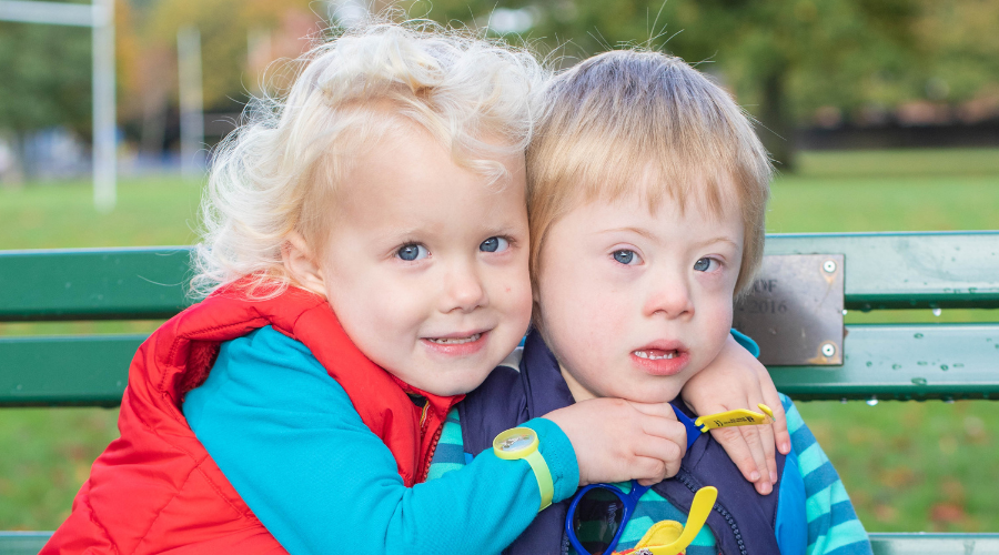 A girl sits on a park bench with her arm round her brother.