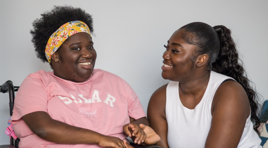 Two adult sisters sit closely, grinning and holding hands. One sister uses a wheelchair.