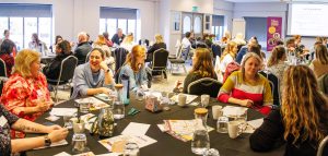 A large room of people sit at round tables, chatting and laughing