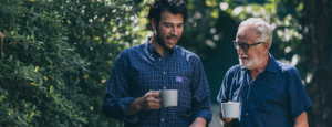 A younger man and an older man have a relaxed walk and talk outdoors, with cups of coffee