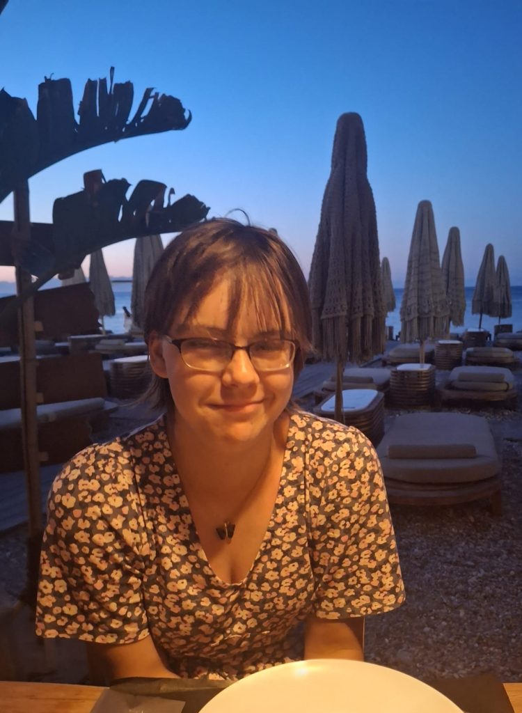 Kathryn faces the camera smiling, wearing glasses and her hair tied back. A beach with sunloungers and palm trees is in the background.