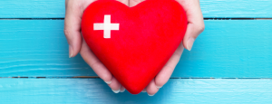 Two hands hold a red heart with a white cross on it; against a blue background