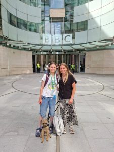 Hannah and her autistic sister Sally stand side by side smiling outside the BBC studios in London. Benji the dog sits at their feet.