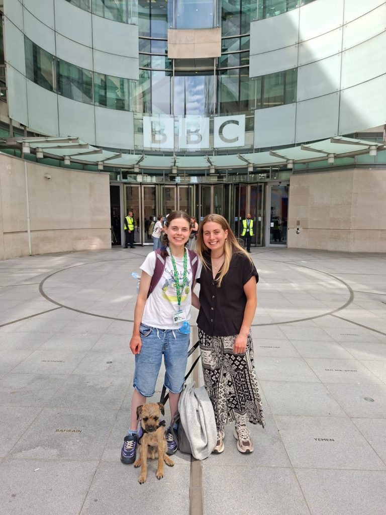 Hannah and her autistic sister Sally stand side by side smiling outside the BBC studios in London. Benji the dog sits at their feet.