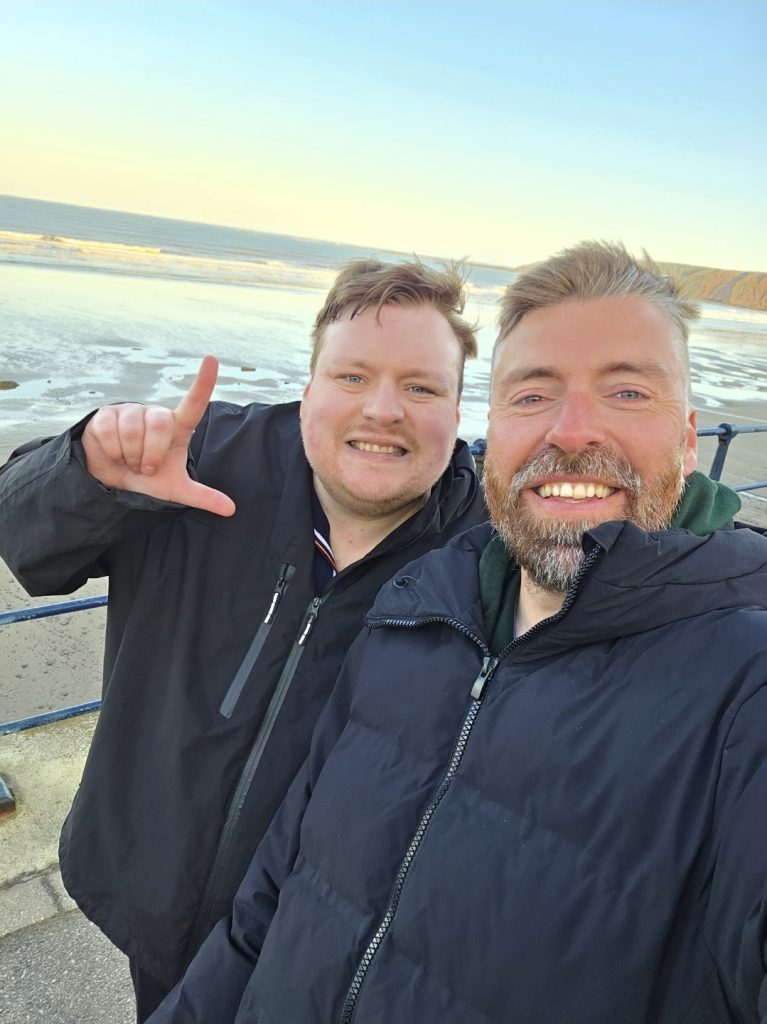 A selfie taken by Adam with his brother on a cool windy day out at the beach. Adam's brother holds up his right hand to make an 'L' sign with his fingers.