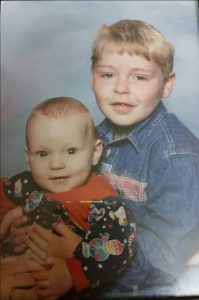 A young Adam sits behind his baby brother, with his arms wrapped around him and they both turn to face the camera