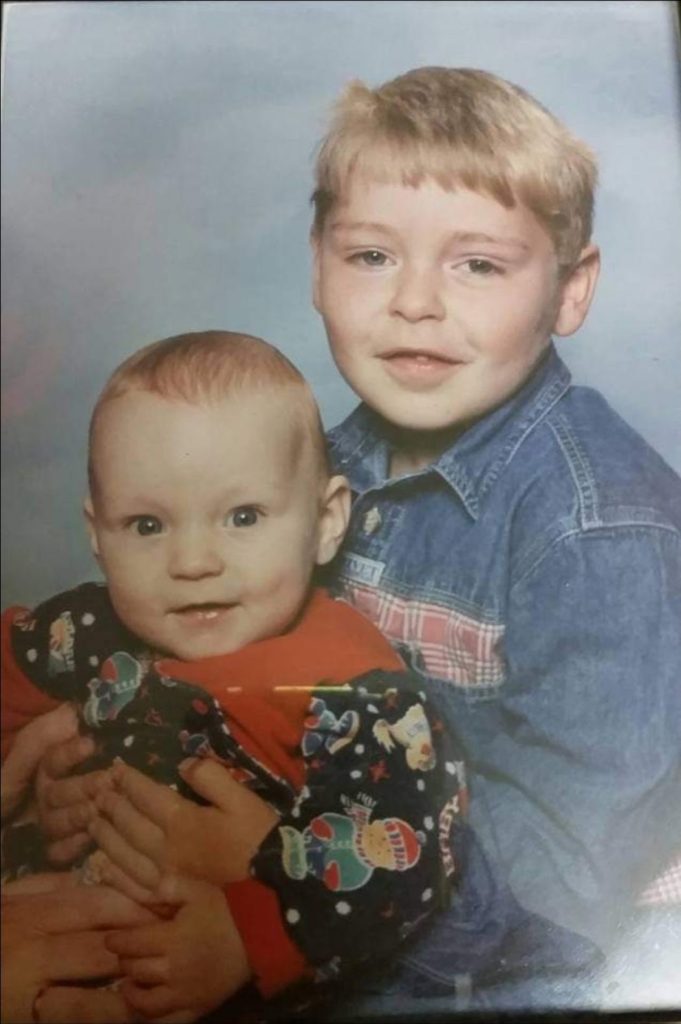 A young Adam sits behind his baby brother, with his arms wrapped around him and they both turn to face the camera
