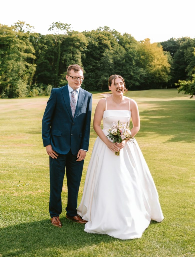 Katie wears a white strapless wedding dress, and holds a bouquet of flowers, she stands next to her brother in a suit. both are smiling, possibly laughing.